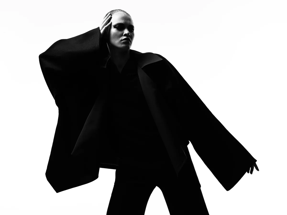 Editorial photograph of a model in a wide sleeved black jacket, silhouette against white